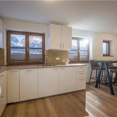 A modern kitchen with wooden floor and large windows. Snow-covered mountains are visible in the background.