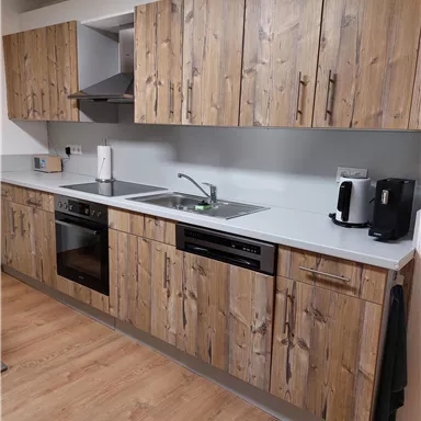 Eine moderne Küche mit Holzfronten und einem klaren, minimalistischen Design. Die Ausstattung umfasst einen Herd, eine Spüle und eine Kaffeemaschine.