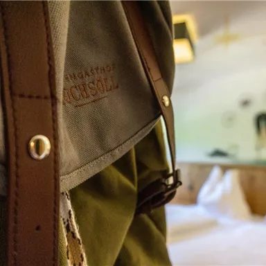 A close-up look at a backpack with leather details. In the background, a cozy room with white beds and a mural can be seen.