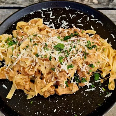 Ein Teller mit Pasta und einer cremigen Sauce, garniert mit frischem Parmesan und Petersilie. Die Nudeln sind auf einem schwarzen Teller angerichtet und auf einer rustikalen Holzoberfläche platziert.