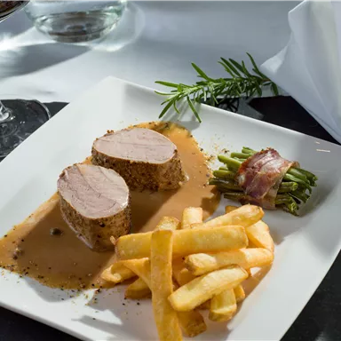 A delicious dish with tender meat, a creamy sauce, and crispy french fries. It is served with fresh green beans wrapped in bacon and a glass of wine in the background.