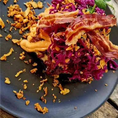 Ein Teller mit lila Krautsalat, knusprigem Speck und Röstzwiebeln. Das Gericht liegt auf einem schwarzen Teller und ist dekorativ angerichtet.