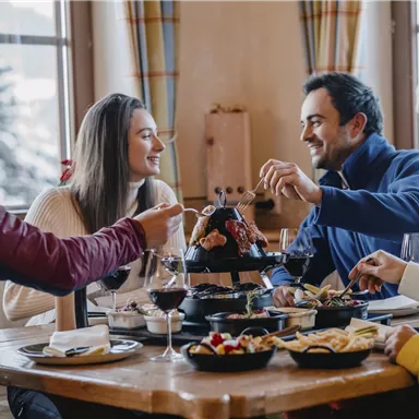 A group of four friends is sitting at a table, enjoying a meal together. The atmosphere is cozy and inviting, with wine and various dishes on the table.