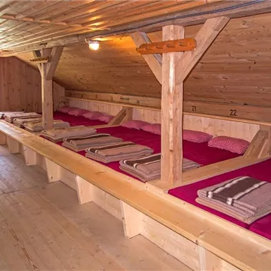 Ein gemütlicher Schlafraum mit mehreren Betten aus Holz. Die Betten sind mit roten Kissen und Decken ausgestattet.