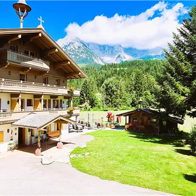 A beautiful chalet in the Alps with lush greenery and tall trees. In the background, impressive mountains and a blue sky can be seen.