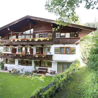 Ein charmantes Haus im alpinen Stil mit mehreren Balkonen und bunten Blumen. Umgeben von üppigem Grün und Bäumen, vermittelt es eine einladende Atmosphäre.