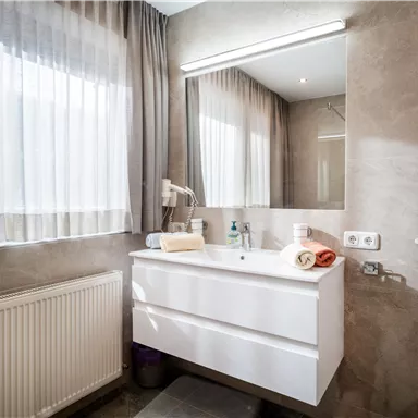 A modern bathroom with a bright vanity and a large mirror. Soft curtains provide plenty of daylight.