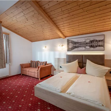 A cozy hotel room with a wooden ceiling and a red carpet. The room features a double bed, a seating area, and a picture on the wall.