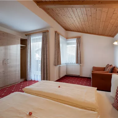 A cozy bedroom with two beds and a wardrobe. The room has large windows and a light wooden ceiling.