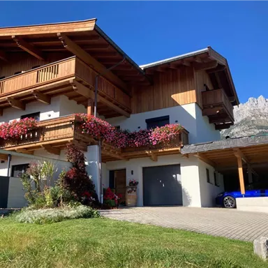 A beautiful wooden house with flower boxes and a spacious entrance. In the background, mountains and a blue sky can be seen.