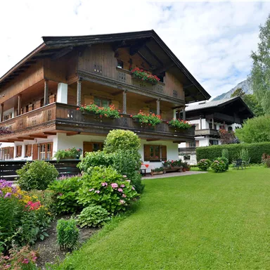 A traditional alpine house with blooming balconies and a well-maintained garden. In the background, mountains and a clear sky can be seen.