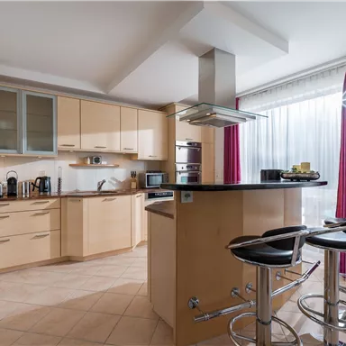 A modern kitchen with wooden cabinets and a central dining area. Large windows let in plenty of light and offer a view of the outdoor area.