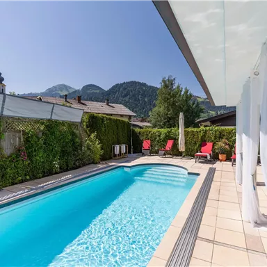 A beautiful outdoor pool with clear water and red loungers. In the background, there are mountains and a green landscape visible.