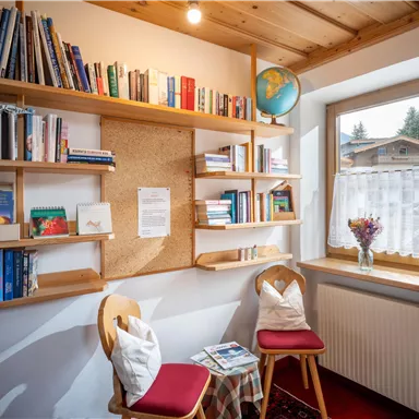 Ein gemütlicher Leseecke mit einem Bücherregal an der Wand. Zwei Stühle und ein kleiner Tisch stehen vor einem Fenster mit Blick nach draußen.