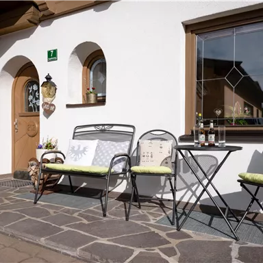 Eine gemütliche Terrasse mit einem Eingang und einem kleinen Tisch. Es gibt zwei bunte Sitzbänke und eine freundliche, einladende Atmosphäre.