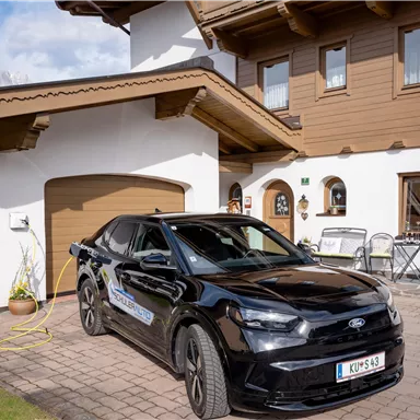 Ein schwarzes Auto steht vor einem traditionellen, holzverkleideten Haus. Im Hintergrund sind einige Gartenmöbel und ein klarer Himmel sichtbar.