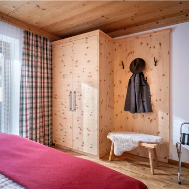 Ein gemütliches Schlafzimmer mit Holzwänden und einem großen Kleiderschrank. Auf einem Bett liegt eine rote Decke und neben steht eine Bank mit einem Hut.