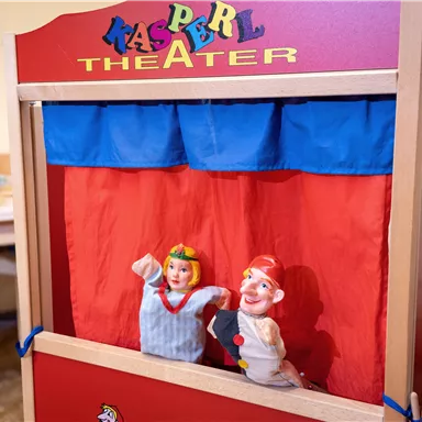Ein Kasperle-Theater mit bunten Puppen steht auf einem Tisch. Die Puppen zeigen einen Kasper und ein Mädchen, die auf eine Aufführung warten.