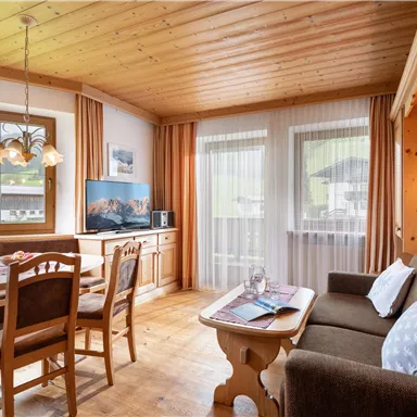 Ein gemütlicher Wohnraum mit Holzdecken und großen Fenstern. Es gibt einen Esstisch, einen Fernseher und eine Sitzecke mit einem Sofa.