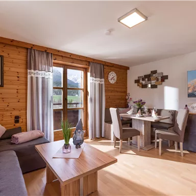 A cozy living kitchen with wooden walls and large windows. The space is bright and inviting, furnished with a dining table and a comfortable couch.
