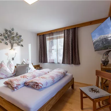 A cozy bedroom with a double bed, nice pillows, and a wooden chair. A television and large windows create a bright, inviting atmosphere.