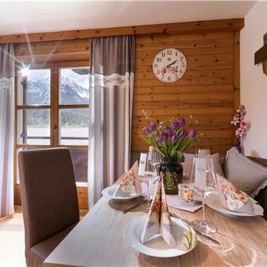 A cozy dining area in a wooden house with a nicely set table. Through the window, you can see the mountains in the background.