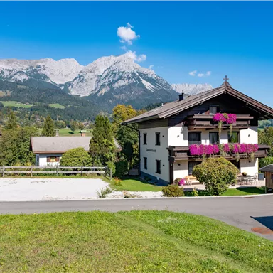 Ein malerisches Haus in einer grünen Umgebung mit blühenden Pflanzen. Im Hintergrund sind hohe Berge unter einem klaren Himmel zu sehen.