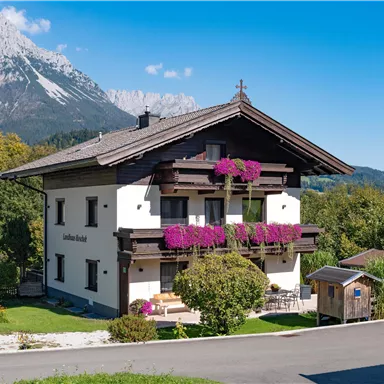 Ein charmantes Haus mit bunten Blumenbalkonen und einem klaren blauen Himmel. Im Hintergrund sind majestätische Berge zu sehen.