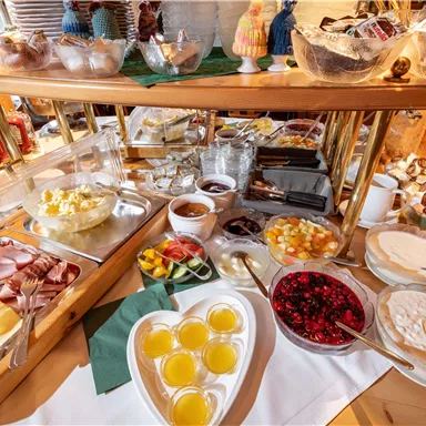 Ein reichhaltiges Frühstücksbuffet mit verschiedenen Käse- und Wurstsorten, frischem Obst und Joghurt. Farbiges Dessert und Säfte ergänzen die Auswahl.
