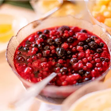 Ein Schüssel mit fruchtiger Beerenmischung, die aus roten und schwarzen Beeren besteht. Im Hintergrund sind weitere Schüsseln mit verschiedenen Obstsorten zu sehen.