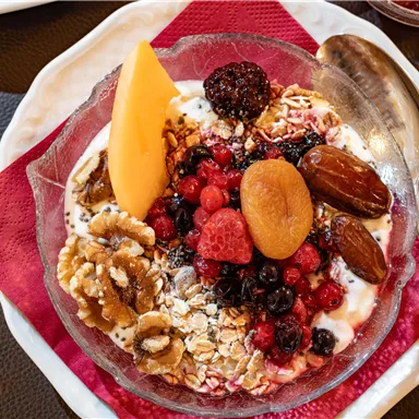 Eine Schüssel mit Joghurt, Müsli und frischen Beeren ist dekoriert mit Nüssen, Datteln und einer Scheibe Melone. Rot und braun dominieren die Farben des gesunden Frühstücks.