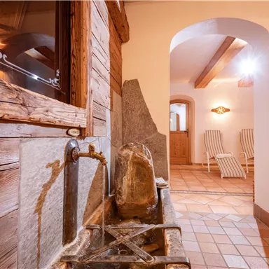Ein gemütlicher Wellnessbereich mit einem traditionellen Brunnen aus Holz und Stein. Im Hintergrund sind Liegen und eine einladende Atmosphäre sichtbar.
