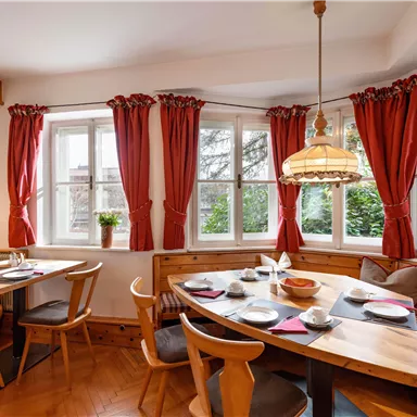 Ein gemütlicher Essbereich mit einem runden Holztisch und bequemen Stühlen. Große Fenster mit roten Vorhängen sorgen für eine einladende Atmosphäre.