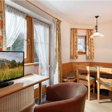 Ein gemütlicher Wohnraum mit einem Fernseher und einem Esstisch aus Holz. Die Fenster sind mit blumigen Vorhängen dekoriert und lassen viel Licht herein.