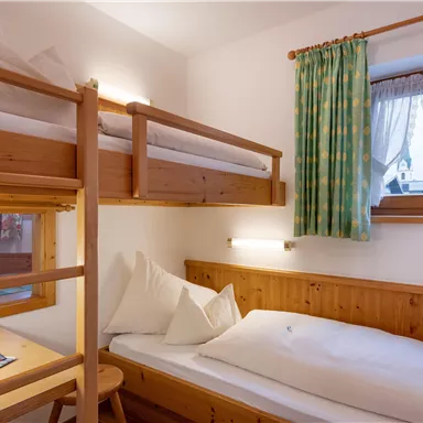 Ein gemütliches Zimmer mit einem Etagenbett und einem Einzelbett. Die Wände sind hell und es gibt ein Fenster mit Vorhängen, das Tageslicht hereinlässt.