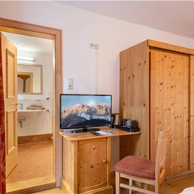 Ein gemütliches Schlafzimmer mit einem Fernseher, einem Holzschrank und einem kleinen Tisch. Im Hintergrund ist das Badezimmer sichtbar.