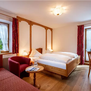 Ein gemütliches Zimmer mit einem Doppelbett und einem roten Sessel. Große Fenster fluten den Raum mit Licht und bieten Ausblick ins Freie.