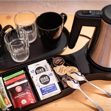Eine Teestation mit einem Wasserkocher, zwei Tassen, Gläsern und einer Auswahl an Teebeuteln und Kaffee. Es sind auch Löffel und Sahne-Behälter vorhanden.