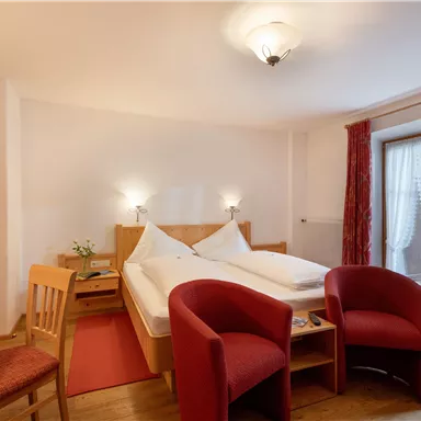 Ein gemütliches Schlafzimmer mit zwei Einzelbetten und roten Sesseln. Fenster mit Vorhängen bringen natürliches Licht in den Raum.