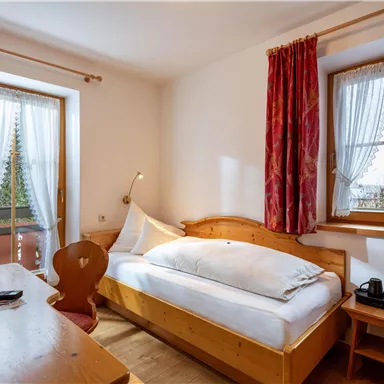 Ein gemütliches Zimmer mit einem Einzelbett, Holzdekor und einem Schreibtisch. Große Fenster lassen viel Tageslicht herein und bieten einen Blick nach draußen.