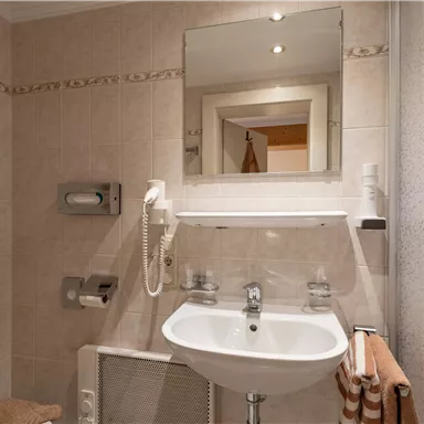 Ein modernes Badezimmer mit Fliesen in creme und einem kleinen Waschbecken. Im Hintergrund ist ein Handtuchhalter und ein Spiegel zu sehen.