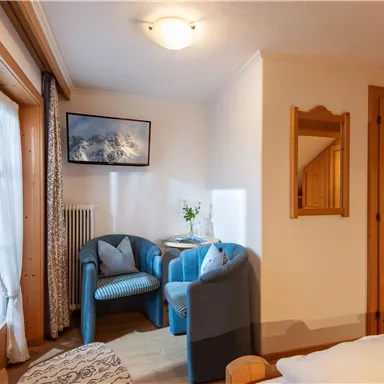 Ein gemütlicher Raum mit blauen Sesseln und einem Tisch. Ein Fenster mit Vorhängen und ein Bild an der Wand ergänzen die Atmosphäre.