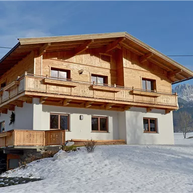 Ein gemütliches Holzhaus mit einem Balkon, umgeben von schneebedecktem Boden. Im Hintergrund sind sanfte Berge zu sehen.