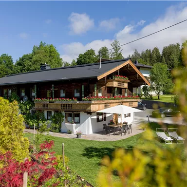 Ein schönes Haus im Grünen mit einem großen Garten. Auf der Terrasse stehen Möbel und Blumen blühen in der Umgebung.
