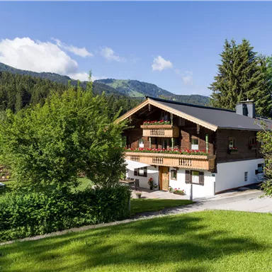 Ein hübsches Chalet umgeben von grünen Wiesen und Bäumen. Im Hintergrund sind Berge und ein blauer Himmel zu sehen.