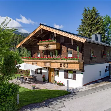 Ein charmantes Holzhaus mit Balkonen und flowerndem Blumenschmuck. Im Vordergrund befindet sich eine Terrasse und im Hintergrund sind Bäume und ein blauer Himmel zu sehen.