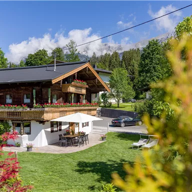 Ein gemütliches Haus mit einem Balkon und einem Garten. Die Umgebung ist grün und idyllisch, umgeben von Bäumen und Blumen.