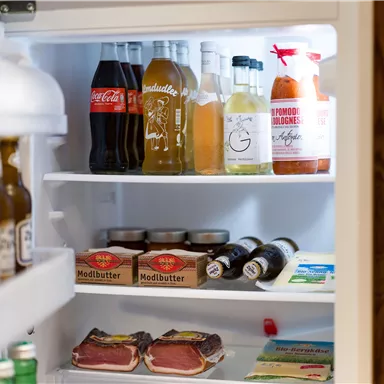 Ein Kühlschrank, der mit verschiedenen Getränken und Lebensmitteln gefüllt ist. Es sind unter anderem Cola, Fleisch und Modlbutter sichtbar.