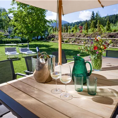 Ein gemütlicher Tisch im Garten, dekoriert mit Gläsern, einer Karaffe und einer Blumenvase. Im Hintergrund sind grüne Bäume und ein gepflegter Garten zu sehen.
