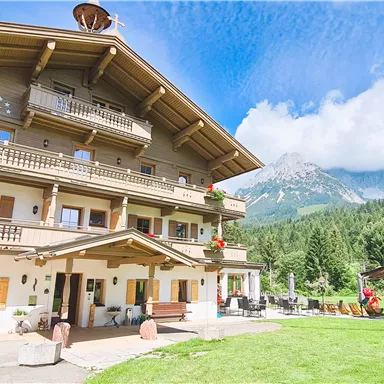A charming Alpine hotel with a balcony and blooming flower boxes. In the background are green meadows and high mountains.